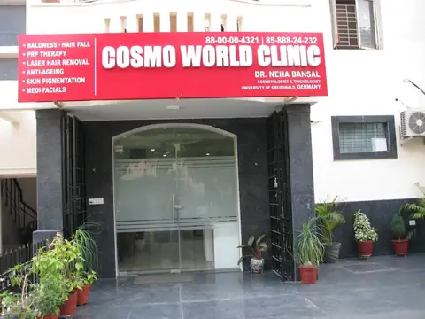 The clinic's ground-floor entrance, with a red signboard reading Cosmo World Clinic and Dr. Neha Bansal above glass double doors, and potted plants lined along the paved forecourt.